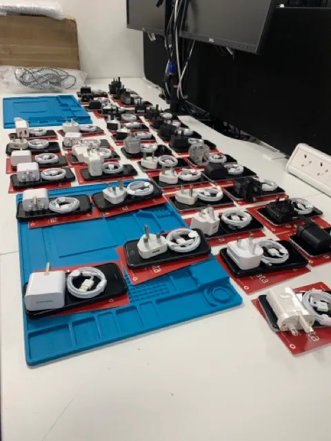 Multiple charging adapters and coiled charging cables organized on red boards placed on blue mats on a white table in front of computer monitors.