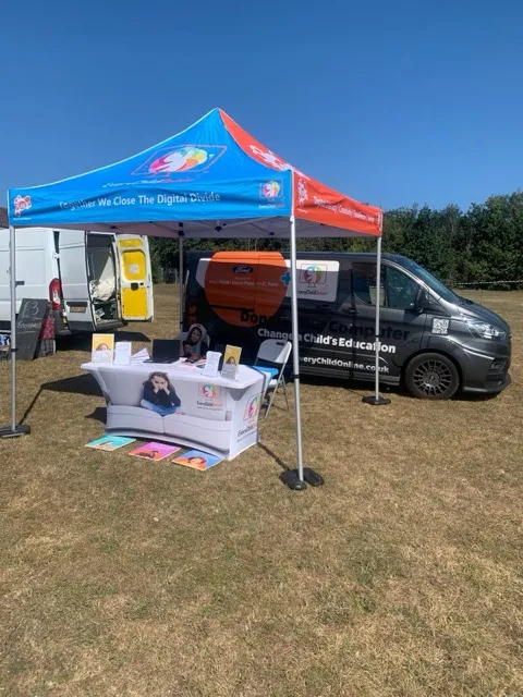 Outdoor awareness booth with canopy tent and table promoting digital divide closure and child education on grass field under clear blue sky.