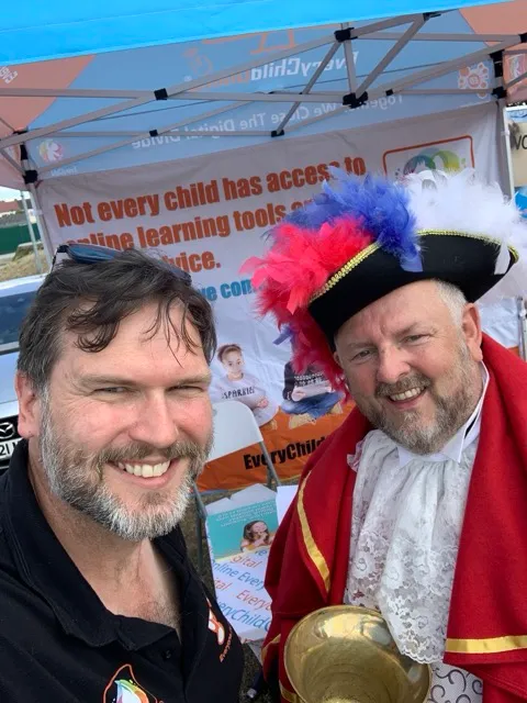 Two men smiling at the camera; one wearing a black shirt, the other dressed in a red and white historical costume with a feathered hat, standing in front of a banner about online learning access.
