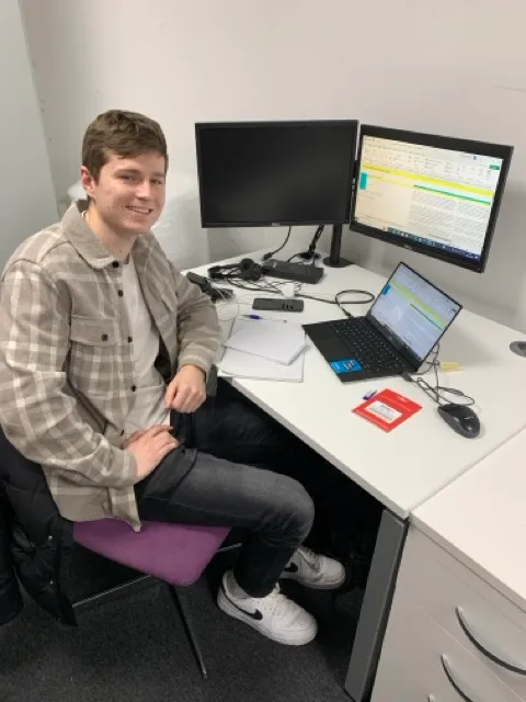 Young man sitting in an office chair at a desk with a laptop, dual monitors, papers, and a computer mouse.