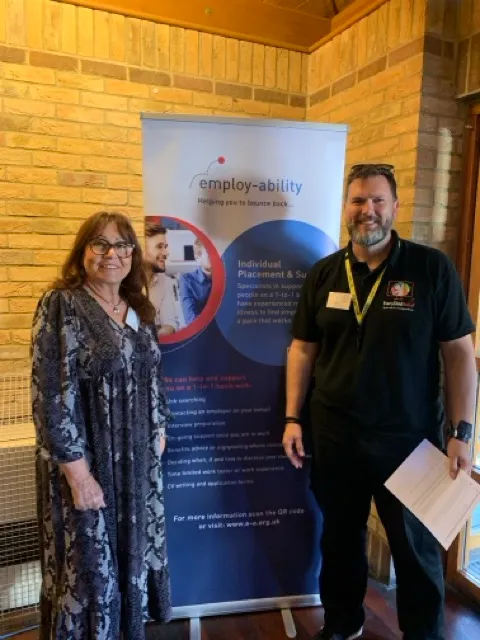 A woman in a patterned dress and a man in a black polo shirt stand beside an Employ-ability information banner in a brick-walled room.