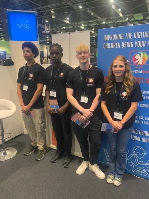 Four young people wearing black t-shirts with lanyards standing in a row at an event booth with educational flyers in their hands.
