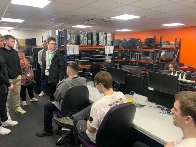 A group of people in a computer lab with several desks, monitors, and shelves holding computer towers and laptops.