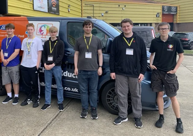 Six young men wearing ID badges standing in front of a dark blue van with company branding outside a yellow building.