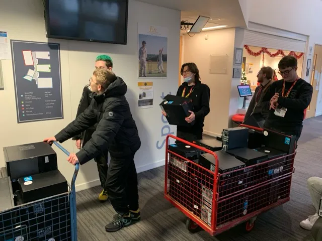 Group of young people moving several computer towers using two red and blue metal carts in an indoor setting.