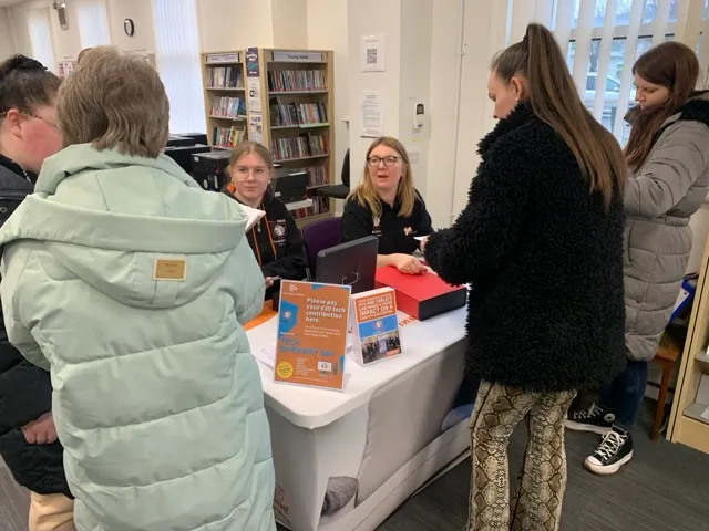 Two women seated behind a table in a library assisting three standing customers, with informational brochures on the table.
