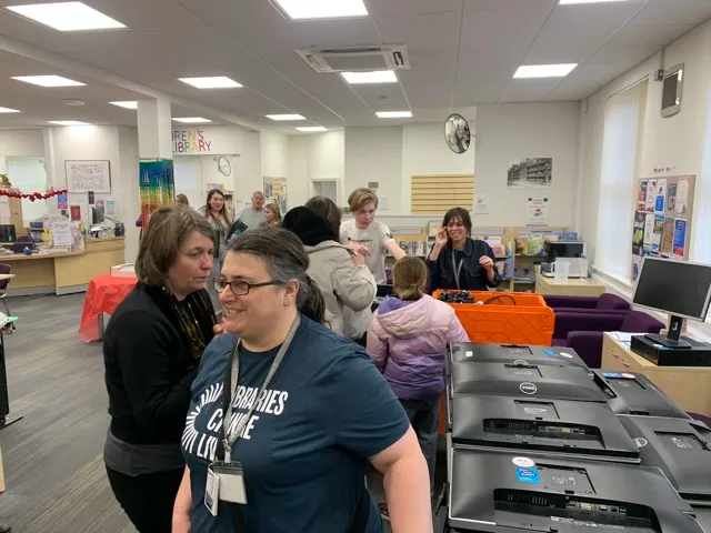 Group of people gathered inside a public library near computer monitors and book shelves.