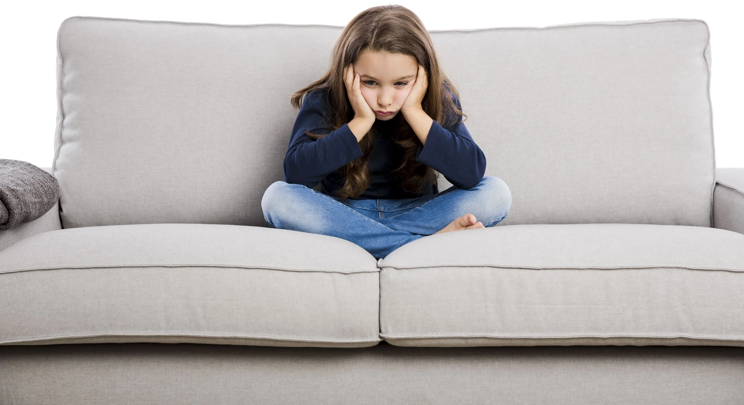 Sad girl sitting cross-legged on a beige sofa with hands on her cheeks.