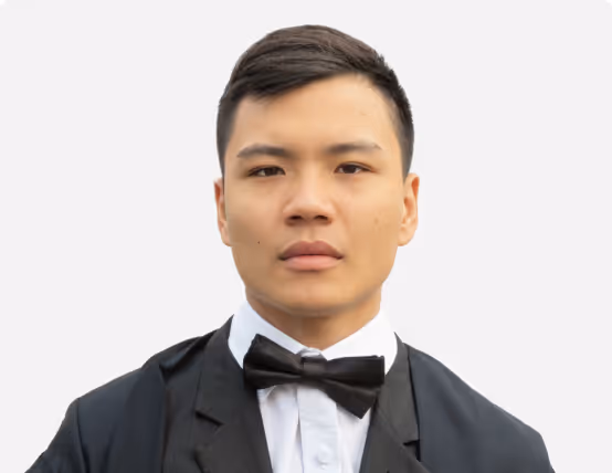 Portrait of a young man wearing a black tuxedo, white shirt, and black bow tie against a plain light background.