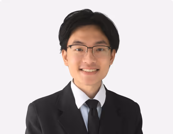 Young man with glasses wearing a black suit, white shirt, and black tie smiling against a plain light background.