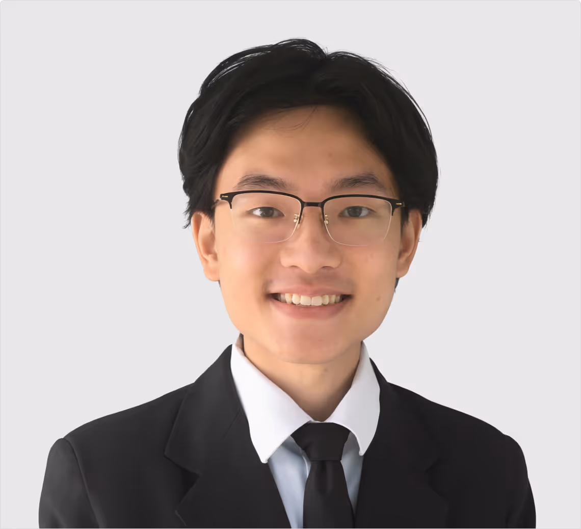 Young man with black hair and glasses wearing a black suit and tie, smiling against a plain light background.
