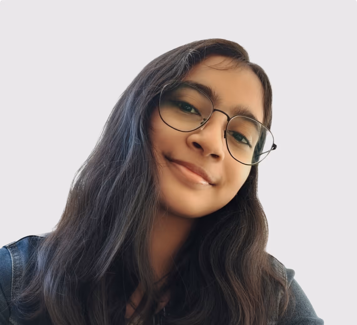 Young woman with long dark hair, wearing glasses and a dark jacket, smiling softly against a light background.