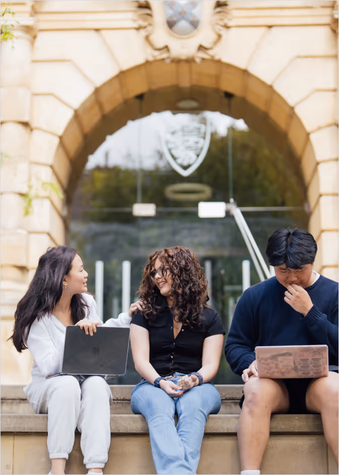 Three college students sitting on steps outside a historic building, two chatting with laptops while the third looks at his device.