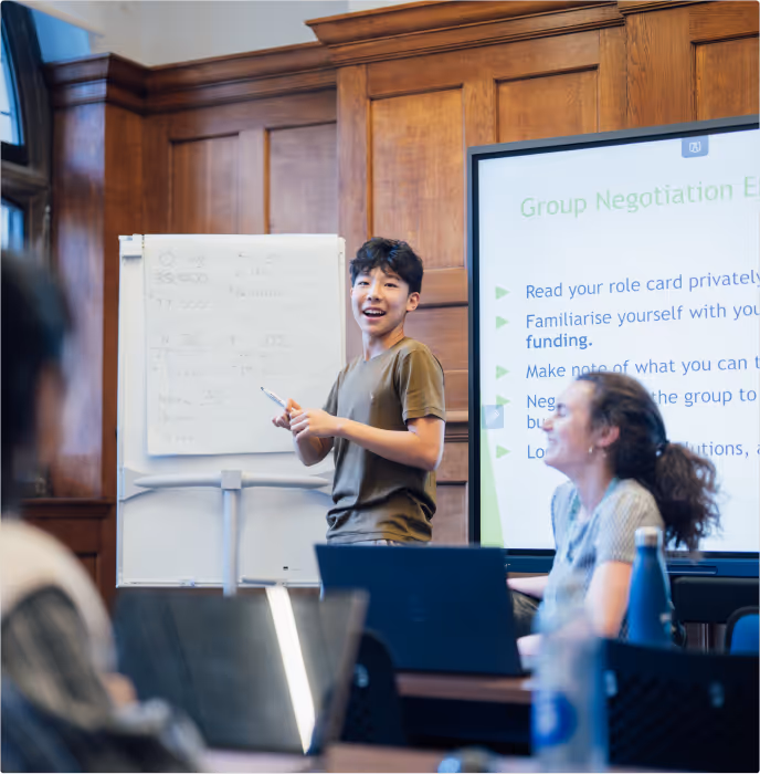 Young person standing and presenting in a classroom with a whiteboard and a screen showing group negotiation instructions.