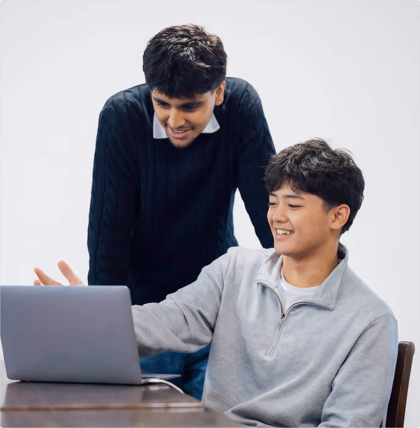 Two young men smiling and looking at a laptop on a wooden table against a white background.