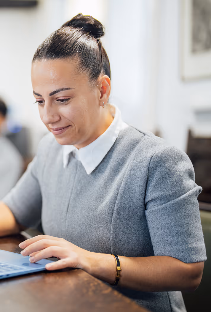 Woman with dark hair tied in a bun wearing a gray dress with white collar, working on a laptop at a wooden table.