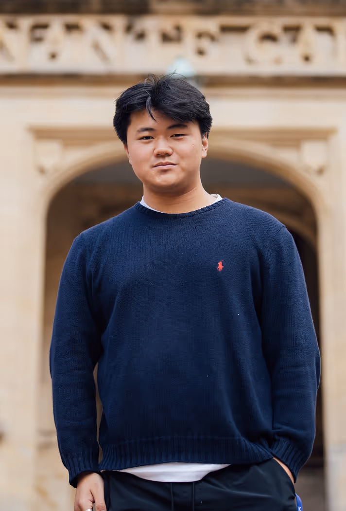 Young man wearing a navy blue sweater standing outdoors in front of a stone archway.