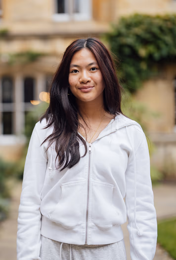 Young woman with long dark hair wearing a white zip-up hoodie standing outdoors in front of a blurred building and greenery.