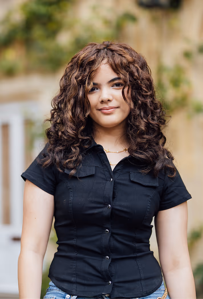 Young woman with curly hair wearing a black button-up shirt and smiling outdoors.