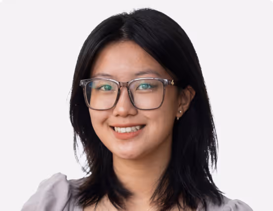Smiling young woman with black hair, wearing large glasses and a light gray top, against a plain white background.