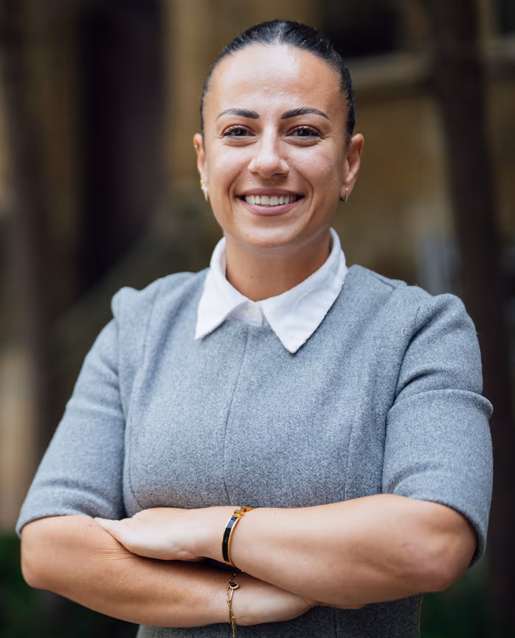 Smiling woman with dark hair pulled back, wearing a gray dress with a white collar and crossing her arms.