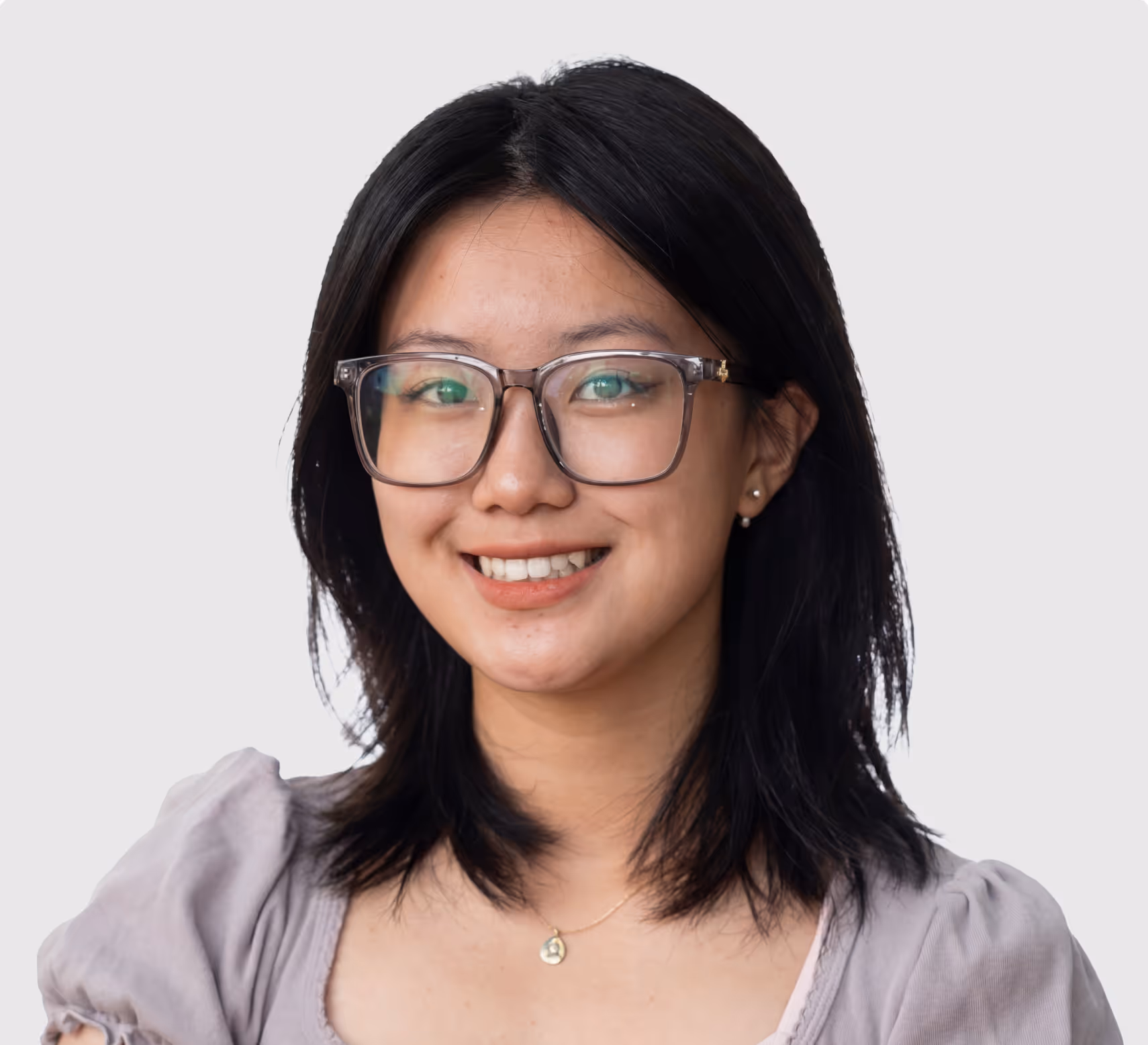 Smiling young woman with black hair, wearing glasses and a light purple top against a plain background.