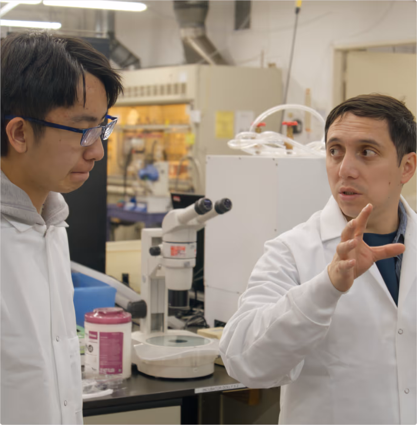 Two scientists in white lab coats discussing in a laboratory with a microscope and lab equipment in the background.