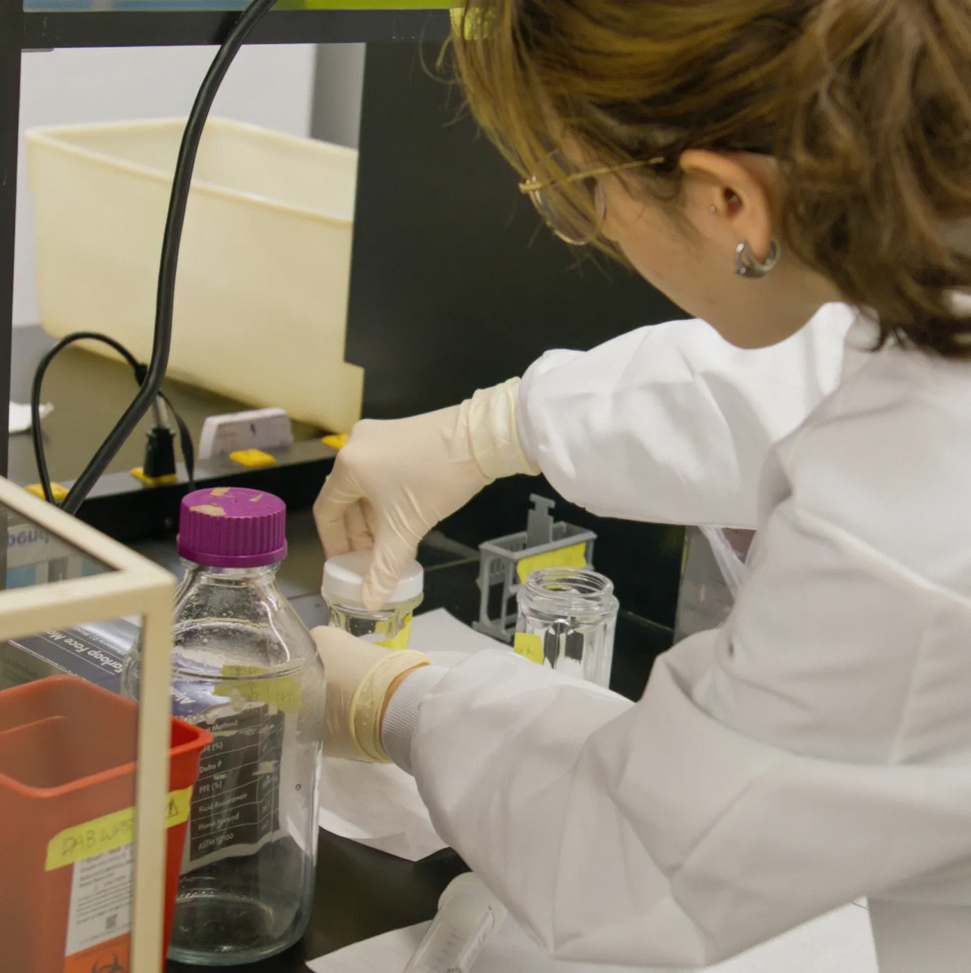 Scientist wearing gloves and a lab coat handling glass containers on a laboratory bench.
