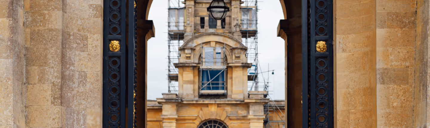 View of a historic stone building with scaffolding through a large archway supported by stone columns and adorned with black and gold decorative metalwork.