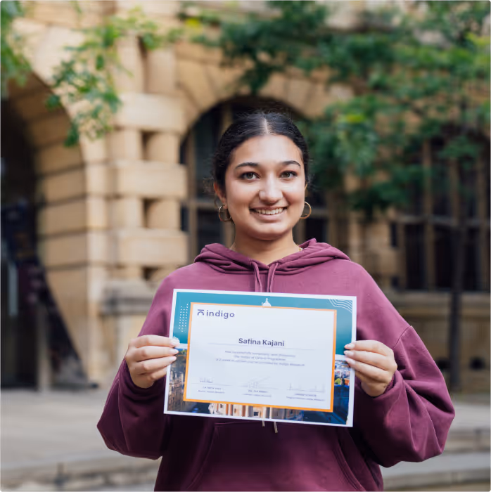 Smiling young woman in a purple hoodie holding a certificate with the name Safina Kajani, standing outdoors in front of a building.