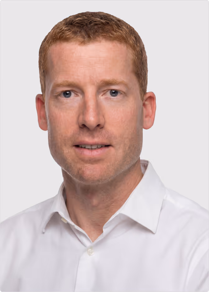 Portrait of a man with short red hair and blue eyes wearing a white collared shirt against a light gray background.