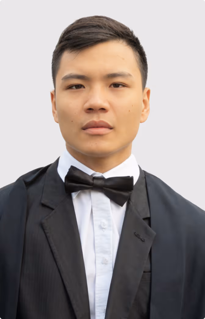Young man wearing a black tuxedo with a white shirt and black bow tie against a plain light gray background.