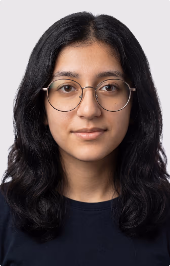 Young woman with long black wavy hair, wearing round glasses and a dark top, looking straight ahead with a neutral expression.