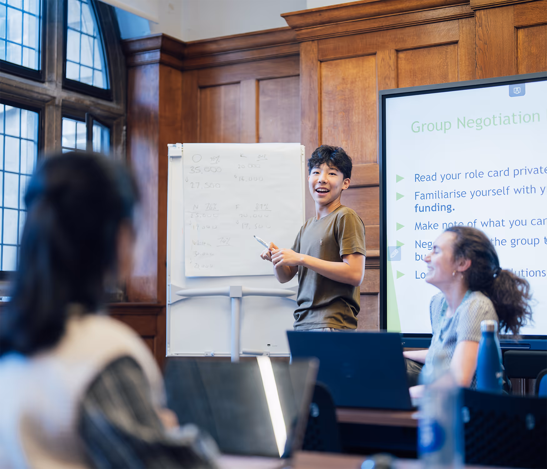 Young man presenting and smiling in a wood-paneled room with a flipchart and a screen showing group negotiation instructions.