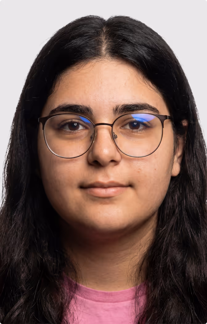 Close-up portrait of a young person with long black hair and round glasses wearing a pink shirt against a gray background.