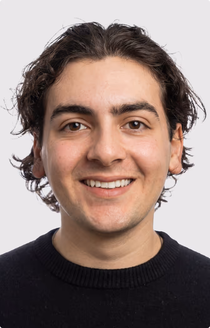 Young man with wavy dark hair, brown eyes, and a black sweater, smiling against a plain white background.