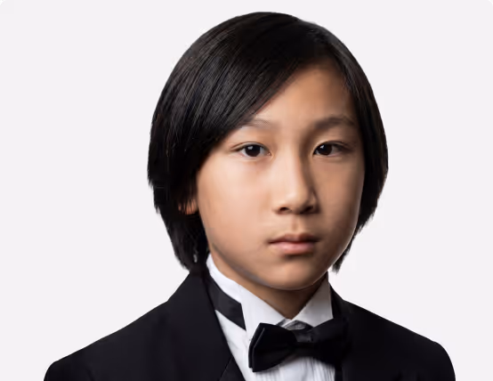 Young boy with straight black hair wearing a black tuxedo and bow tie against a white background.