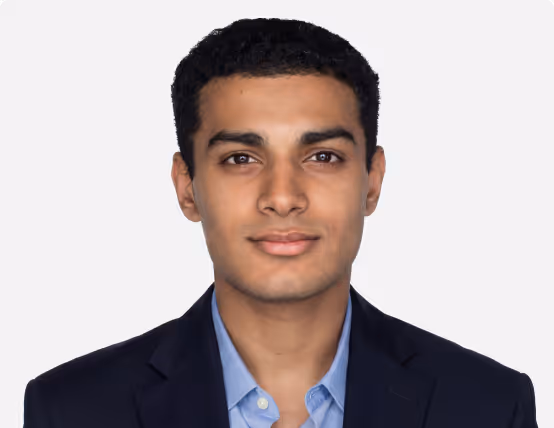 Portrait of a young man with short curly black hair wearing a navy blazer and light blue shirt.
