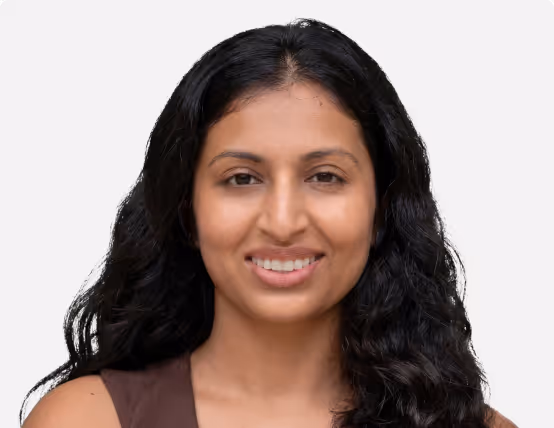 Smiling woman with long curly black hair wearing a sleeveless brown top against a plain light background.