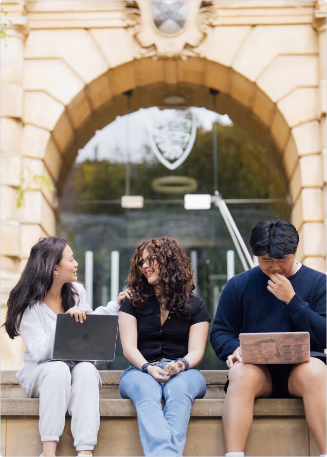 Three young adults sitting on stone steps outside a building, two women smiling and talking, one man focused on a laptop.