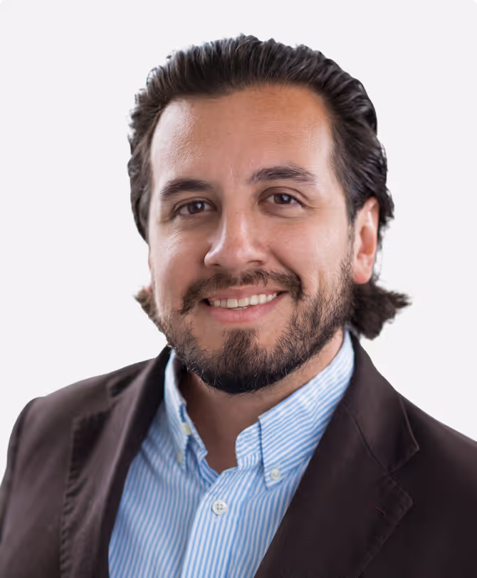 Smiling man with dark hair and beard wearing a brown blazer and blue striped shirt.
