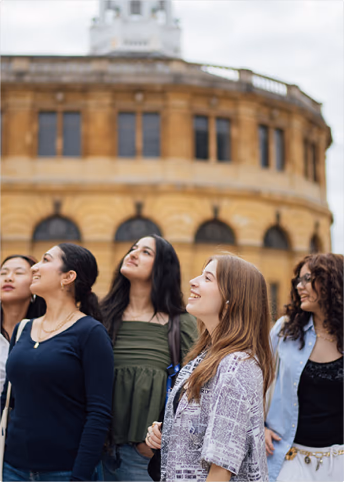 Group of young people standing and talking on a courtyard lawn in front of an old stone building with multiple windows.