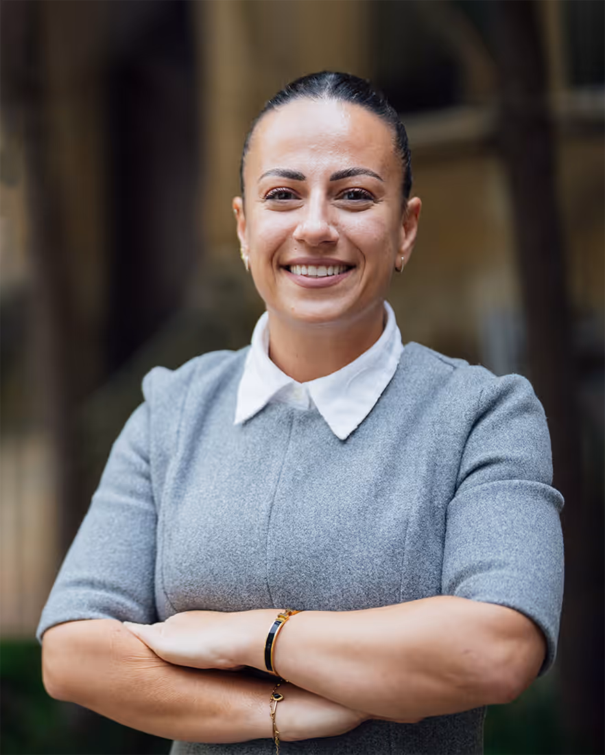 Smiling woman with dark hair pulled back, wearing a gray dress with a white collar and crossing her arms.