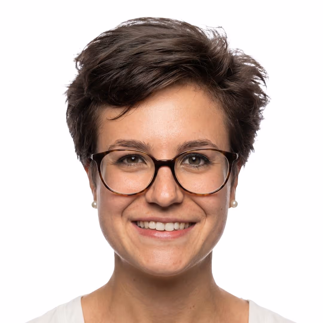 Smiling woman with short brown hair, wearing tortoiseshell glasses and pearl earrings, against a white background.