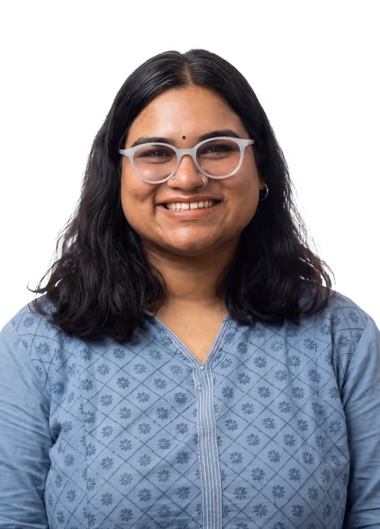 Smiling woman with glasses, nose ring, and dark wavy hair wearing a blue patterned top.