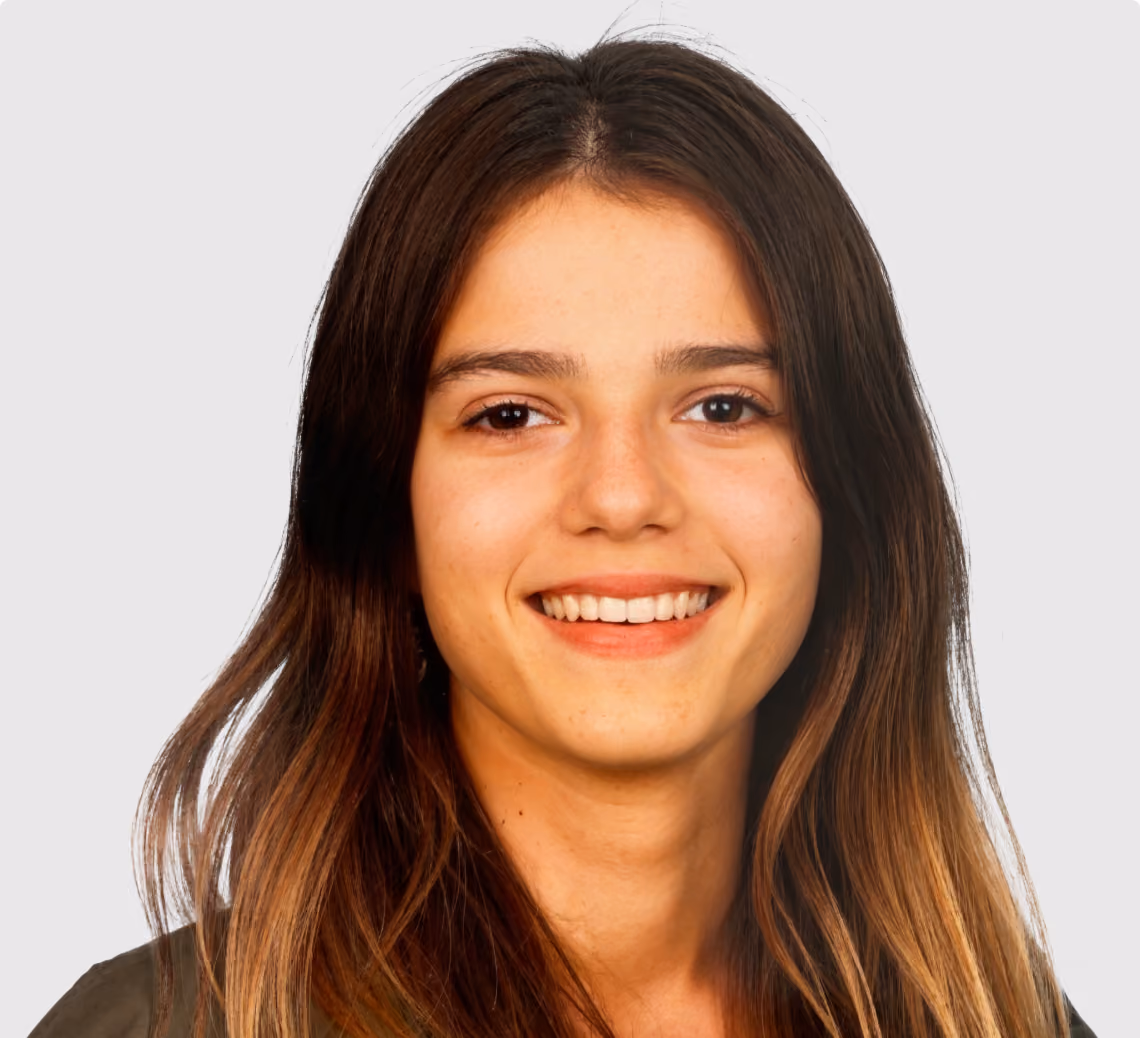 Young woman with long brown hair smiling in front of a plain light gray background.