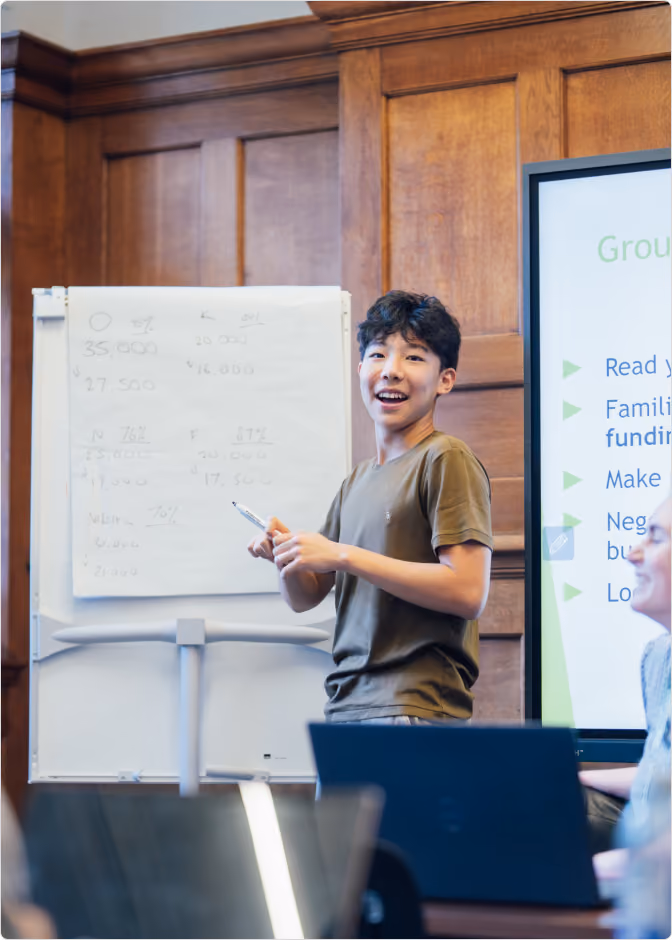 Young man standing and smiling while presenting in a classroom with a flip chart and a screen showing group negotiation exercise instructions.
