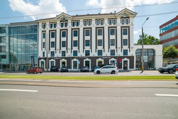 Street view of a large white and black building with multiple windows and cars parked in front on a sunny day.