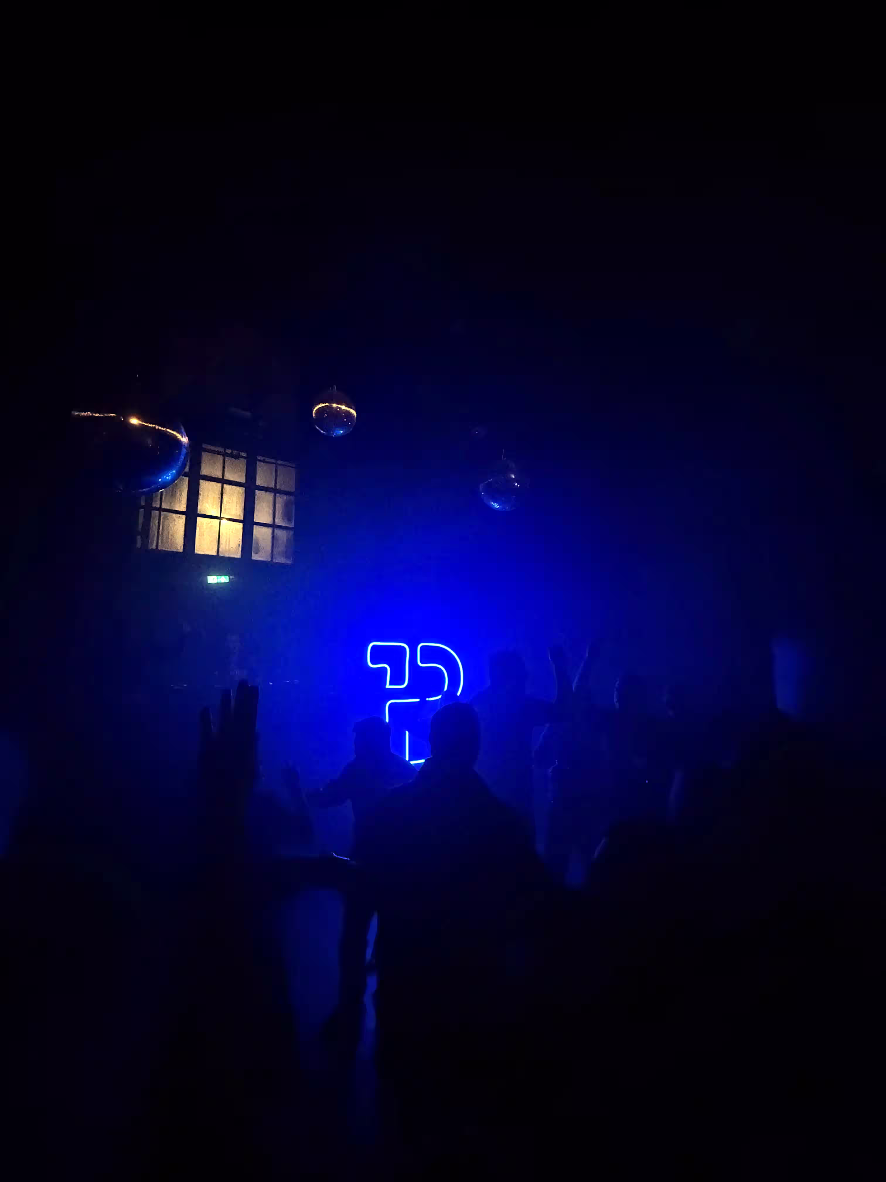 Silhouettes of people dancing in a dark venue, lit by a glowing blue neon Backbase logo on the wall, with mirror balls overhead.