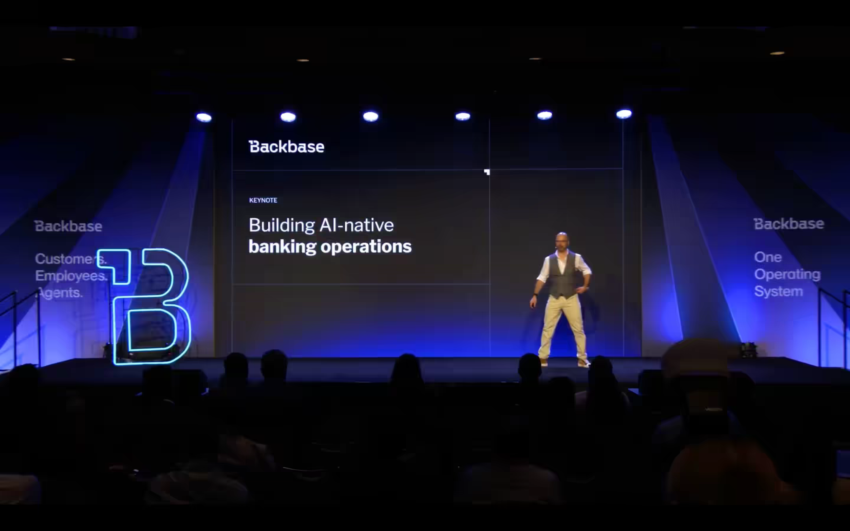 A presenter on stage at a Backbase Engage event delivering a keynote titled "Building AI-native banking operations." The Backbase logo and branded stage elements are visible, along with side panels reading "Customers. Employees. Agents." and "One Operating System."